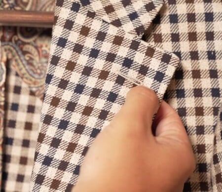 A hand indicating the lapel buttonhole on a beige, navy, and brown checked jacket, shown up close